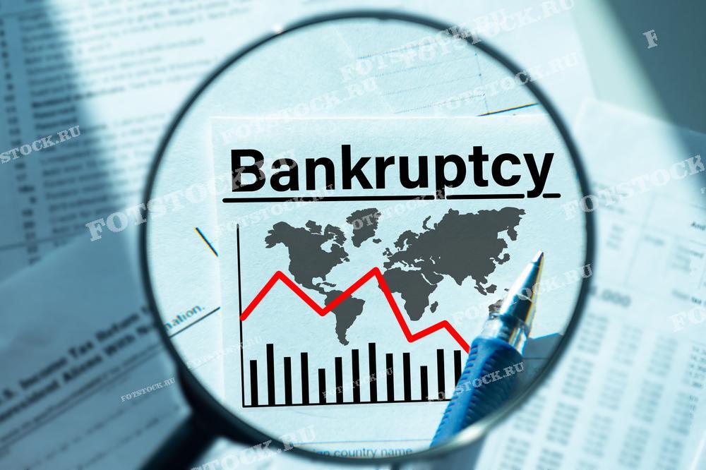 На фотографии изображены документы с графиком падения и текстом "Bankruptcy" под лупой. На графике видна карта мира, символизирующая глобальный экономический спад и распространение финансовых проблем.